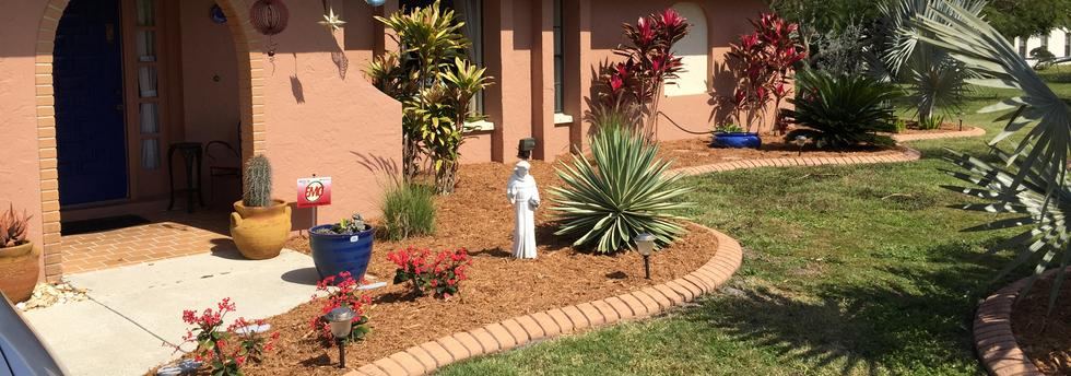 Brick-edged bed of bromeliads and shrubs running along a coral home's front lawn
